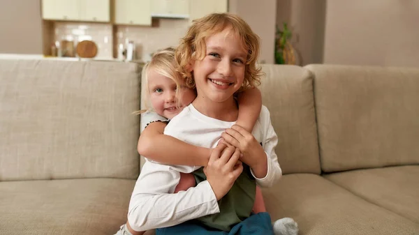 Портрет радостных братьев и сестер, мальчик и девочка, улыбающиеся в камеру, обнимающие друг друга, сидя дома на диване — стоковое фото