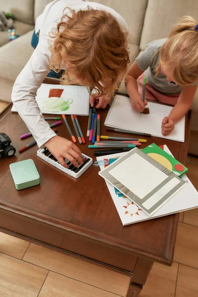 Beskuren bild av upptagna små barn, pojke och flicka rita på papper med markeringspenna, sitter tillsammans på en soffa hemma — Stockfoto