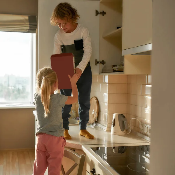 Retrato de menino impertinente encontrado cornflakes caixa no armário da cozinha e dando-o a sua irmãzinha — Fotografia de Stock