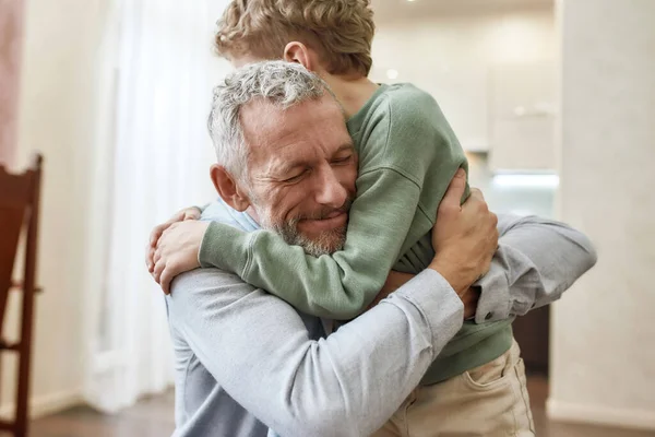 Емоційна зустріч. Щасливий дідусь обіймає свого милого маленького дошкільного онука, стоячи разом вдома — стокове фото