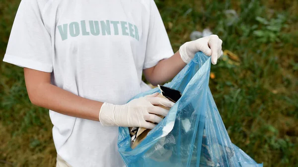 Takarítási nap. Levágott kép egy fiatal önkéntesről, aki egyenruhát és gumikesztyűt visel szemeteszsákkal a kezében, miközben az erdőt vagy a parkot takarítja a szemétből. — Stock Fotó