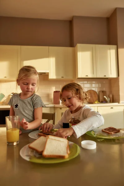 Küçük kız kardeşi ve kendisi için mutfakta kahvaltı hazırlarken tatlı erkek kardeş tost ekmeğine çikolata ezmesi sürüyor. — Stok fotoğraf