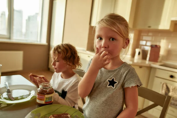 Küçük sevimli bir kızın portresi. Kameraya bakıyor, çikolatalı tereyağı tadıyor ve kardeşiyle birlikte mutfakta kahvaltı ediyor. — Stok fotoğraf