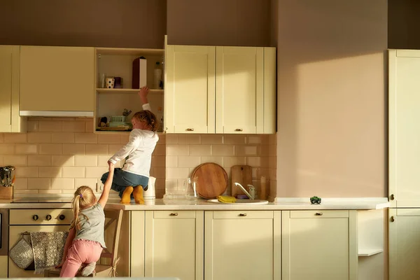 Vue arrière de deux vilains enfants, frère et sœur essayant de trouver quelque chose de doux dans l'armoire de cuisine. Petite fille aidant son frère en lui tenant la main — Photo