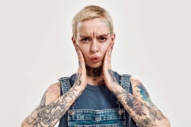 Duygular ve duruşlar. Beyaz piercingli ve dövmeli bir kadın kameraya bakıyor. Kot pantolon giyiyor. Elleri yanaklarında sevimli yüz ifadeleri sergiliyor.
