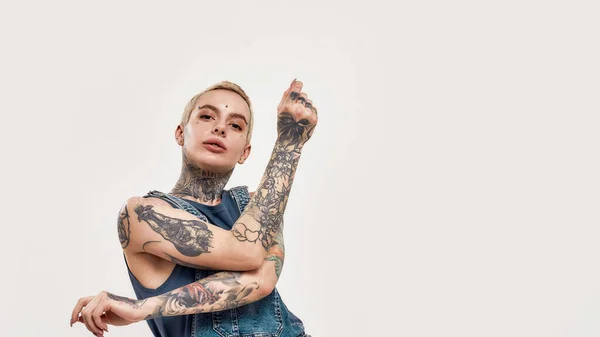 Tatuagem e piercing. Uma mulher tatuada branca com piercing dançando sozinha usando uma ganga geral olhando para uma câmera com as mãos cruzadas em um movimento — Fotografia de Stock
