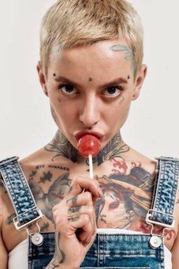 Dövme ve piercing. Dövmeleri olan, kot pantolon giyen, kameraya bakarken lolipop emen beyaz bir kadının yakın çekimi.