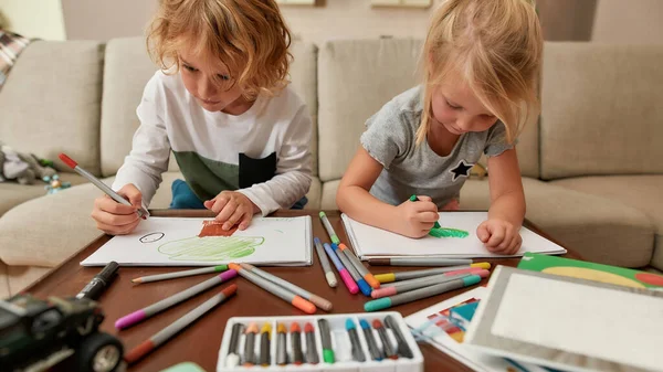 Odaklanmış küçük kardeşler, kağıt üzerine çizim yapan kız ve erkek kardeşler evde birlikte vakit geçirirken kalem kullanıyorlar. — Stok fotoğraf