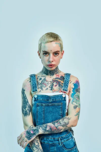 Tatuaje y piercing. Una chica blanca tatuada con piercing vistiendo denim en general de pie con las manos cruzadas sobre su vientre mirando a una cámara — Foto de Stock