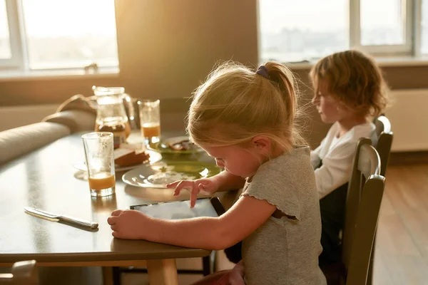 Retrato de menina bonita focada usando tablet pc enquanto toma café da manhã ou almoço, sentado junto com seu irmão na mesa na cozinha — Fotografia de Stock