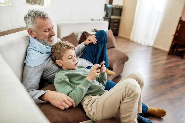 子供や近代的な技術。幸せな祖父と自宅でソファの上でリラックスしながら、スマートフォンを使用して小さな男の子孫 — ストック写真