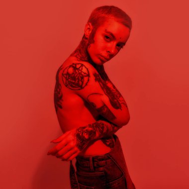 Dövme ve piercing. Dövmeleri ve piercingleri olan beyaz bir kadın elleri göğsünde, göğüslerini kapatan bir kameraya bakıyor.