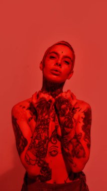 Dövme ve piercing. Beyaz dövmeli, piercingli, üstsüz bir kadın. Elleriyle göğüslerini kapatan bir kameraya bakıyor.