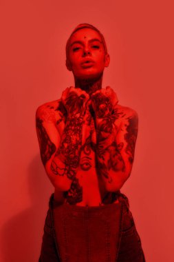 Dövme ve piercing. Dövmeli, piercingli, kot pantolon giyen, göğüslerini kapatan ve kameraya bakan beyaz bir kadın.