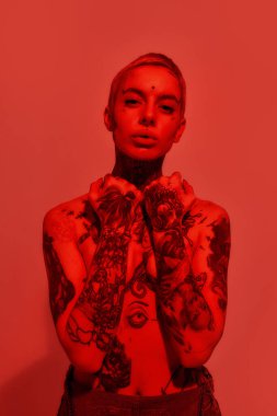 Dövme ve piercing. Dövmeli ve piercingli beyaz bir kadının portresi. Üstsüz duruyor. Elleriyle göğüslerini kapatan bir kameraya bakıyor.