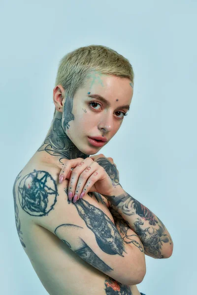 Tattoo and piercing. A close up of a white pierced and tattooed woman standing topless sideways to a camera with her eyes open and hands crossed on her chest covering breasts — Stock Photo, Image