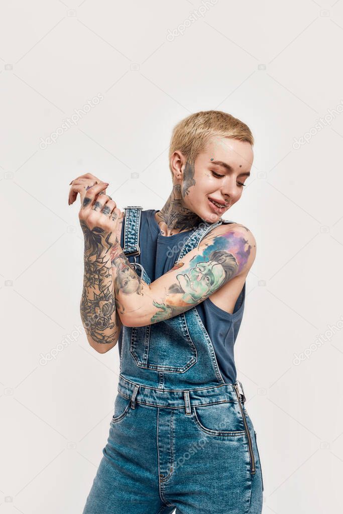 Tattoo and piercing. A white tattooed pierced woman holding her hands together while smiling and dancing wearing a denim overall