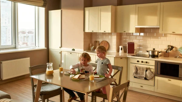 Petit frère et petite sœur déjeunant seuls le matin à la maison. Adorable garçon faisant un sandwich pour lui-même — Photo