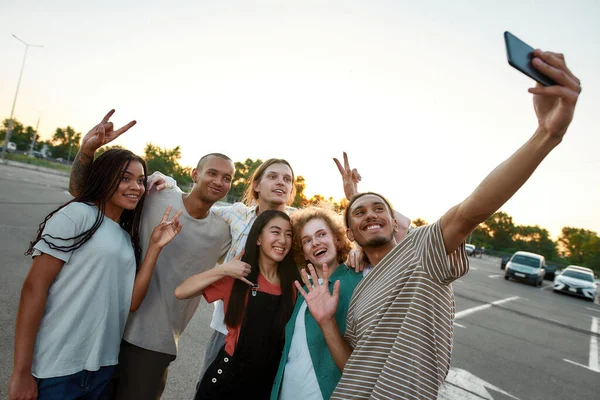 Seis jóvenes amigos bien vestidos pasándola bien juntos abrazándose sonriendo y haciendo un selfie grupal afuera en un sitio de estacionamiento con autos en el fondo —  Fotos de Stock