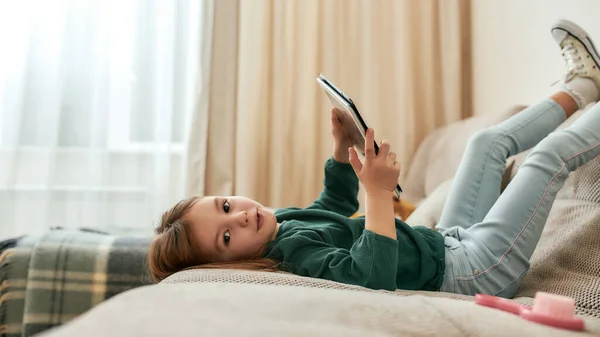 Una piccola ragazza carina che tiene un tablet sdraiato su un divano con i piedi alzati e le gengive sorridenti e guardando in una fotocamera — Foto Stock