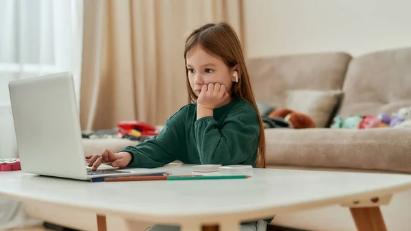 Uma menina pequena bonito sentado sozinho em uma mesa olhando em uma tela de laptop fazendo sua tarefa de educação à distância em uma grande sala brilhante — Fotografia de Stock
