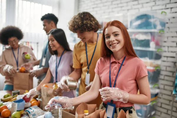 Jonge vrouwelijke vrijwilliger glimlachend op camera tijdens het verpakken van voedsel en dranken donatie in papieren zakken en doos, Kleine groep mensen die werken in liefdadigheidsstichting — Stockfoto