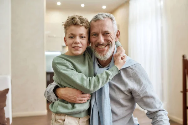 Mutlu büyükbaba, tatlı küçük anaokulu torunuyla kucaklaşıyor, evde birlikte duruyor ve kameraya gülümsüyor. — Stok fotoğraf
