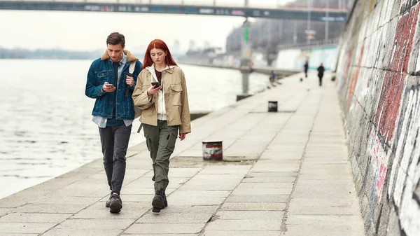 Full length πλάνο των δύο εφήβων απορροφάται εντελώς στα smartphones τους, αγνοώντας ο ένας τον άλλο, ενώ περπατώντας κατά μήκος του ποταμού μαζί — Φωτογραφία Αρχείου