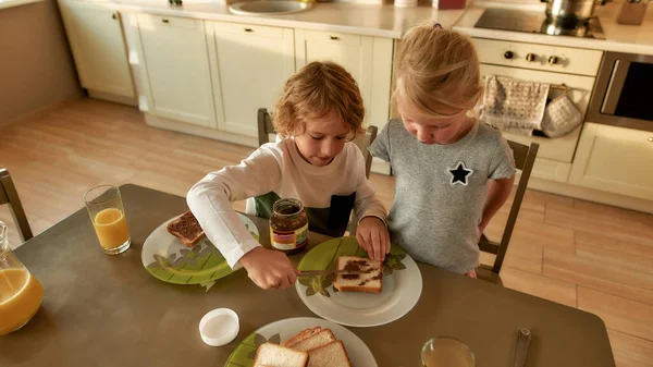 Sevgili ağabeyi, küçük kız kardeşine mutfakta kahvaltı hazırlarken kızarmış ekmeğe çikolata fındıklı tereyağı sürmeyi öğretiyor. — Stok fotoğraf