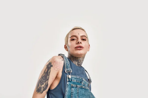 Tetování a piercing. Detailní záběr na bílou tetovanou a propíchnutou ženu, jak se dívá do kamery v džínové kombinéze — Stock fotografie