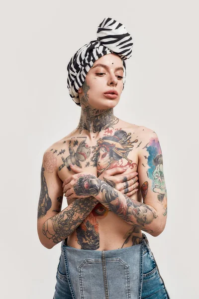 Joven mujer perforada y tatuada de pie en topless con un turbante sobre su cabeza cubriéndose los pechos con las manos cruzadas en el pecho — Foto de Stock
