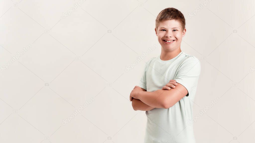 Joyful disabled boy with Down syndrome smiling at camera while posing, standing with arms crossed isolated over white background