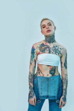 Dövme ve piercing. Beyaz bir kadın dikilirken ve üzerinde beyaz bir bluz olan kot pantolon giyerken kameraya bakan piercing ve dövmeler yaptırıyor.