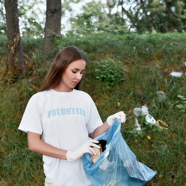 Concepto de voluntariado. Mujer joven, eco activista con uniforme y guantes de goma con bolsa de basura en las manos limpiando bosque o parque de residuos plásticos y basura — Foto de Stock
