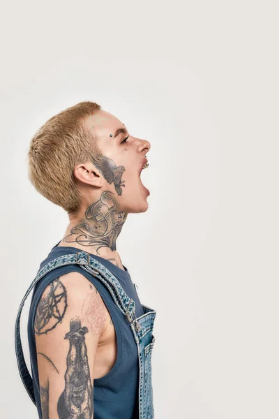Érzelmek és testtartások. Egy fehér, piercinges és tetovált nő, aki oldalt áll a kamera előtt, farmernadrágot visel, és hangosan sikoltozik nyitott szájjal. — Stock Fotó
