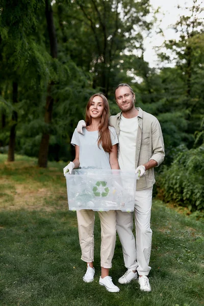 Captura vertical de la joven pareja feliz, eco voluntarios sosteniendo la papelera de reciclaje y mirando a la cámara mientras recogen residuos de plástico juntos en el bosque verde — Foto de Stock