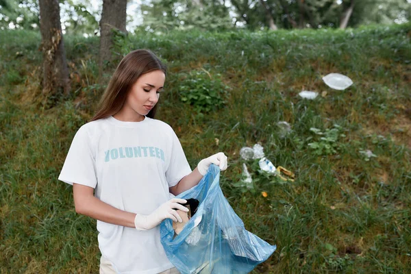 Salva nuestra naturaleza. Mujer joven, eco activista con uniforme y guantes de goma con bolsa de basura en las manos limpiando bosque o parque de residuos plásticos y basura — Foto de Stock