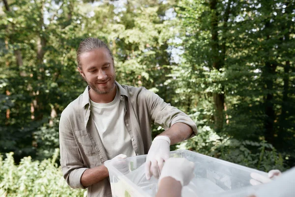 Молодий щасливий чоловік екологічний волонтер, який збирає пластикові відходи з другом в лісі або парку, кидаючи порожні пляшки в кошик для переробки — стокове фото