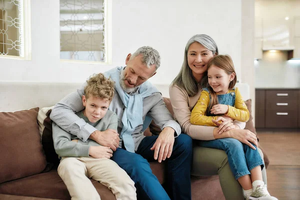 Бабушка и дедушка с внуками обнимаются и улыбаются, сидя вместе на диване в гостиной дома — стоковое фото