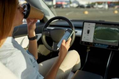 Genç beyaz kız şoför arabanın içinde akıllı telefon kaydırıyor.