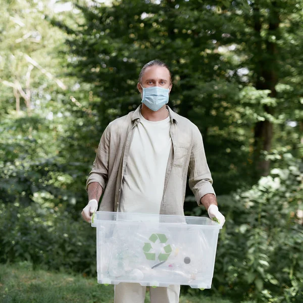 Young caucasian man eco activist wearing protective face mask holding recycle bin and looking at camera while cleaning in the forest during covid 19 outbreak — Stock Photo, Image