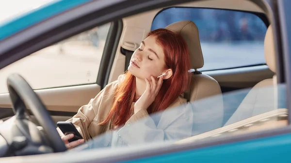 Nastolatka w słuchawkach siedząca w samochodzie z zamkniętymi oczami podczas korzystania z telefonu, słuchająca muzyki — Zdjęcie stockowe