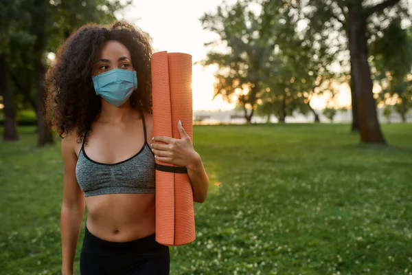 Mantenerse en forma durante el brote de covidio 19. Joven mujer fitness de raza mixta con mascarilla protectora sosteniendo alfombra de yoga y mirando a un lado mientras hace ejercicio en el parque verde —  Fotos de Stock