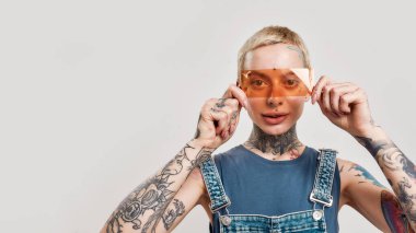 Dövme ve piercing. Üzerinde piercing olan beyaz bir kız. Durmuş, kameraya bakıyor. Elinde turuncu bir filtre var ve gülümsüyor.