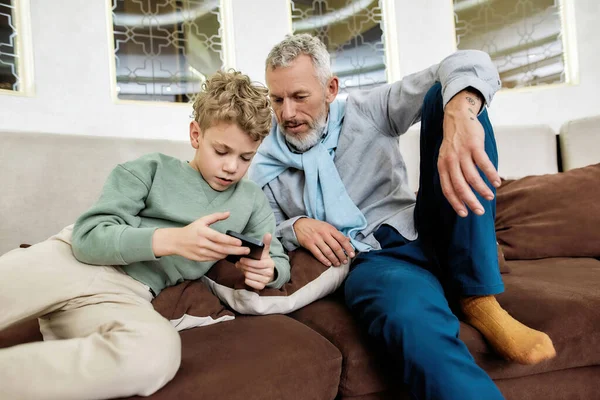 Nonno e il suo nipotino carino seduti insieme sul divano e guardare video o giocare ai videogiochi su smartphone, trascorrere del tempo a hom — Foto Stock