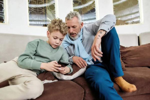 Schattig jongetje tonen zijn grootvader iets op mobiele telefoon, terwijl ontspannen op de bank in het moderne appartement — Stockfoto