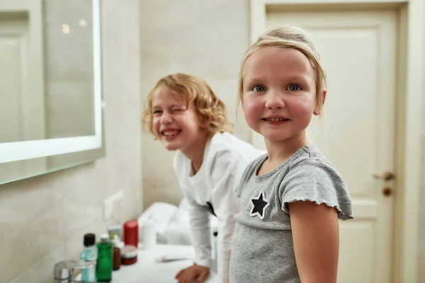 화장실에서 동생과 함께 양치질을 하면서 카메라 앞에서 웃고 있는 귀여운 어린 소녀의 모습 — 스톡 사진