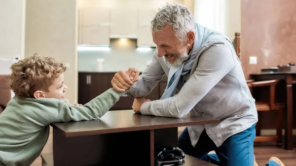 Die sterker is. Gelukkig grootvader en kleinzoon zitten tegenover elkaar en spelen armworstelen, plezier hebben thuis — Stockfoto