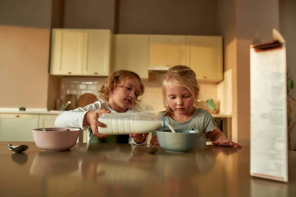 Vårdande bror häller mjölk i en skål flingor medan du förbereder frukost, sitter tillsammans med sin syster vid bordet i köket — Stockfoto