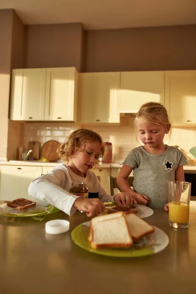 Küçük kız kardeşi ve kendisi için mutfakta kahvaltı hazırlarken tatlı erkek kardeş tost ekmeğine çikolata ezmesi sürüyor. — Stok fotoğraf